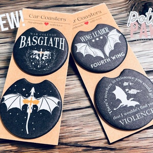 May include: Four black round car coasters with white dragon designs. The coasters feature text including "War College Basgiath", "Wing Leader Fourth Wing", and "There's nowhere in existence you could go that I wouldn't find you, Violence".