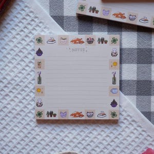 Puede incluir: Un bloc de notas blanco con un borde marrón claro y un interior rayado. El borde está decorado con ilustraciones de gatos, plantas, libros y otros objetos caprichosos. El bloc de notas tiene la palabra "NOTES" escrita en el centro.