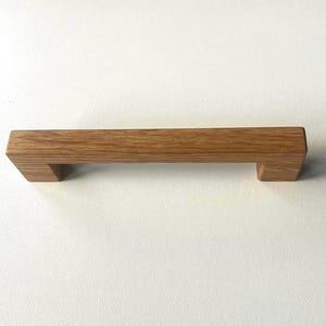 Könnte beinhalten: Ein rechteckiger Möbelgriff aus hellbraunem Holz mit sichtbarer natürlicher Maserung. Der Griff hat ein gerades, horizontales Stangendesign mit zwei kurzen, senkrechten Stützen, die an beiden Enden nach unten verlaufen.