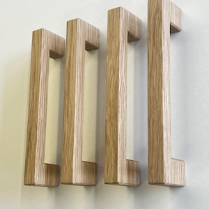 Könnte beinhalten: Vier rechteckige Schrankgriffe aus hellem Holz mit sichtbarer vertikaler Maserung. Jeder Griff hat ein gerades, längliches Design mit zwei rechtwinkligen Befestigungspunkten oben und unten, wodurch ein U-förmiges Profil entsteht.
