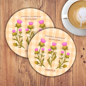 May include: Two round coasters with a checkered pattern in shades of peach and white. The coasters feature a stylised illustration of the thistle, the national flower of Scotland. The text "National Flower of Scotland" and "Thistle" are printed on the coasters.