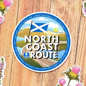 以下が含まれることがあります： 「NORTH COAST ROUTE」の文字とスコットランドの国旗のデザインが施された青と白の丸いステッカー。ステッカーには、道路と山が背景に描かれています。