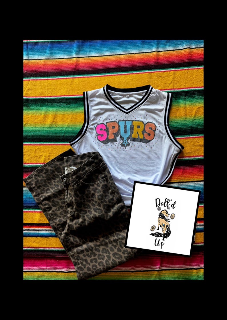 Pu&ograve; includere: Una canotta bianca con scollo a V e bordi neri che riporta la scritta SPURS in lettere colorate, abbinata a pantaloni marroni e neri con stampa leopardata e un cartoncino quadrato con il testo Doll'd Up e l'illustrazione di un cavallo.