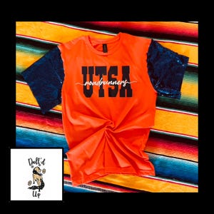 May include: An orange t-shirt with navy blue sequin sleeves. The shirt features the text "UTSA roadrunners" in black. The shirt is displayed on a colorful striped blanket, with a small graphic in the corner.