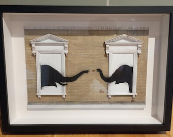 May include: Framed artwork featuring two black dinosaur silhouettes reaching towards each other across a brick wall. The dinosaurs are framed within white window-like structures, creating a unique and artistic composition. The frame is black.