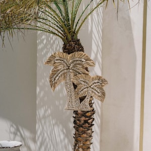 Op de afbeelding: Twee geweven palmboom wanddecoraties die aan een palmboom in een witte pot hangen. De palmboom staat in een kamer met witte muren en witte potten.