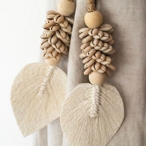 May include: Two beige tassel tiebacks with wooden beads and shell accents. The tassels are made of woven cotton and have a leaf shape. The tiebacks are attached to a beige fabric background.