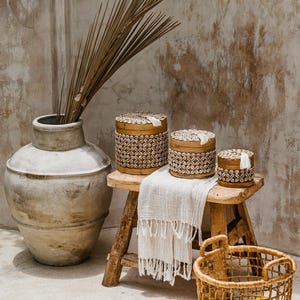 May include: A still life arrangement with a large, weathered ceramic vase holding dried palm fronds. Three decorative wooden boxes with shell and geometric patterns are on a small wooden stool. A woven basket contains lemons.