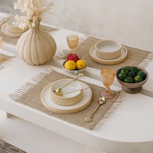 May include: A dining table setting with beige ceramic bowls, plates, and gold-colored spoons. The table is decorated with a beige vase, fruit, and woven placemats. A woven light fixture hangs above the table.