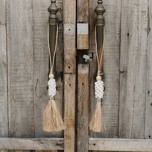 Può includere: Primo piano di una porta in legno usurato con due maniglie in bronzo ornate. Ogni maniglia ha una corda decorativa, un gruppo di conchiglie bianche, una perla di legno e un nappa di fibra naturale. La texture rustica della porta è visibile.