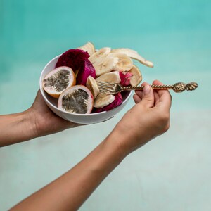 May include: A white bowl filled with a fruit salad of sliced dragon fruit, passion fruit, and banana. A gold pineapple-shaped fork is held in the right hand of a person holding the bowl.