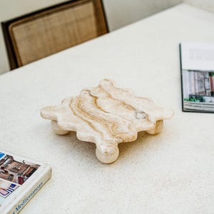 Pode incluir: Uma bandeja decorativa de mármore bege com um design de borda exclusivo e ondulado. A bandeja é suportada por três pés arredondados. O mármore tem veios castanhos naturais. A bandeja está sobre uma superfície branca, com um livro e uma revista por perto.
