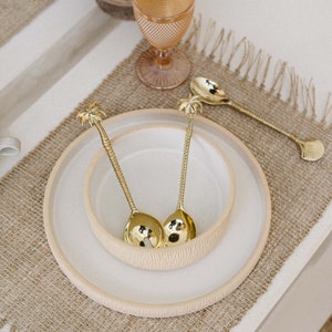 Könnte beinhalten: Zwei goldene Servierlöffel in Palmenform auf einem weißen Keramikteller mit einer kleineren weißen Keramikschale im Inneren. Teller und Schale befinden sich auf einem braunen Jute-Platzdeckchen.