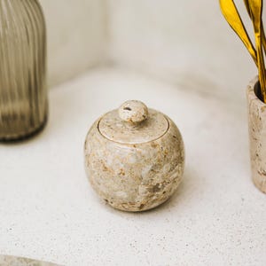 Könnte beinhalten: Ein kleiner, runder Behälter mit Deckel aus beigem Stein mit braunen und beigen Adern. Der Behälter hat einen kleinen, abgerundeten Knopf auf dem Deckel. Goldfarbene Utensilien befinden sich in einem passenden Steingefäß.
