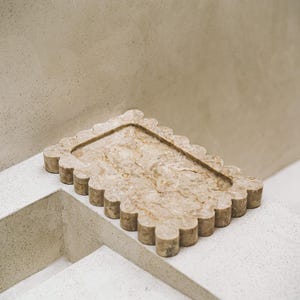 May include: A decorative rectangular tray with a scalloped edge, crafted from beige marble. The tray features a recessed interior and is placed on a white surface, showcasing its natural veining and texture.
