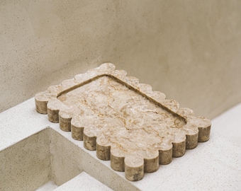 May include: A decorative rectangular tray with a scalloped edge, crafted from beige marble. The tray features a recessed interior and is placed on a white surface, showcasing its natural veining and texture.