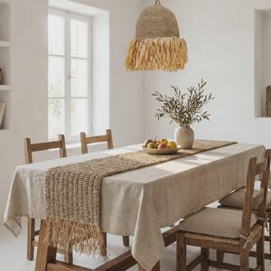 Peut inclure: Un ensemble de salle à manger comprenant une nappe en lin beige, un chemin de table en jute tressé avec des extrémités à franges et des chaises en bois avec des coussins beiges. Un vase en céramique avec des branches d'olivier et un bol de fruits.