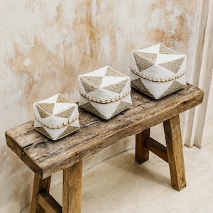 Könnte beinhalten: Drei geometrische Aufbewahrungsboxen in Weiß und Braun mit Perlenbesatz stehen auf einer rustikalen Holzbank. Die Boxen sind nach Größe geordnet, die größte hinten und die kleinste vorne.