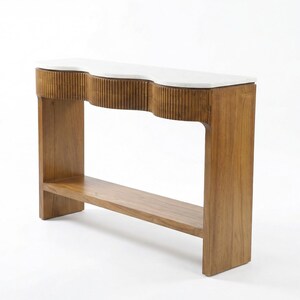 Op de afbeelding: Een houten console met een witte, golvende bovenkant. De tafel heeft een onderste plank en verticale groeven aan de voorkant. Het hout heeft een warme, natuurlijke tint en het ontwerp is modern. De tafel is ongeveer 150 cm lang.