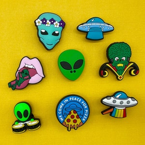 May include: Eight colorful alien-themed shoe charms, including a green alien with a flower crown, a green alien with a tongue sticking out, a green alien playing drums, a green alien with three eyes, a pink mouth with a green alien inside, a blue flying saucer, a green alien with a crown and a green alien with a red collar.