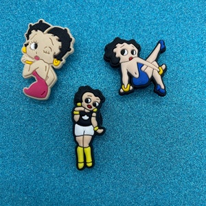 May include: Three cartoon character shoe charms featuring a woman with black hair and a pink dress, a woman with black hair and a blue dress, and a woman with black hair and a black and white outfit.