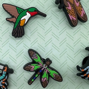 May include: A set of five colorful, detailed, plastic charms featuring a hummingbird, dragonfly, cicada, and two crabs. The charms are on a green background with a chevron pattern.