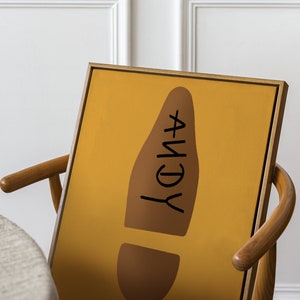 May include: A framed art print featuring a mustard yellow background with a brown footprint shape. The word "ANDY" is written in black within the footprint. The artwork is displayed on a wooden chair.