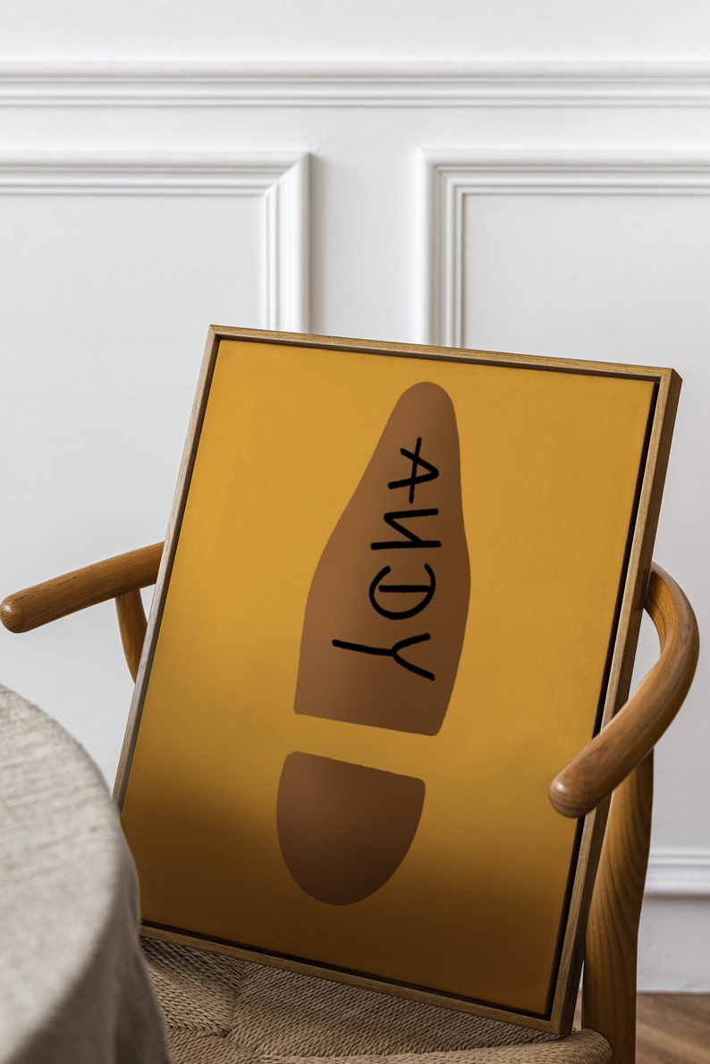 May include: A framed art print featuring a mustard yellow background with a brown footprint shape. The word "ANDY" is written in black within the footprint. The artwork is displayed on a wooden chair.