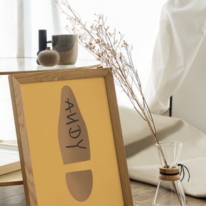 May include: A framed print with a mustard-yellow background features a brown footprint design with the name "ANDY" in black. A glass carafe with a wooden collar and dried flowers are also in the image. The frame is made of light-colored wood.