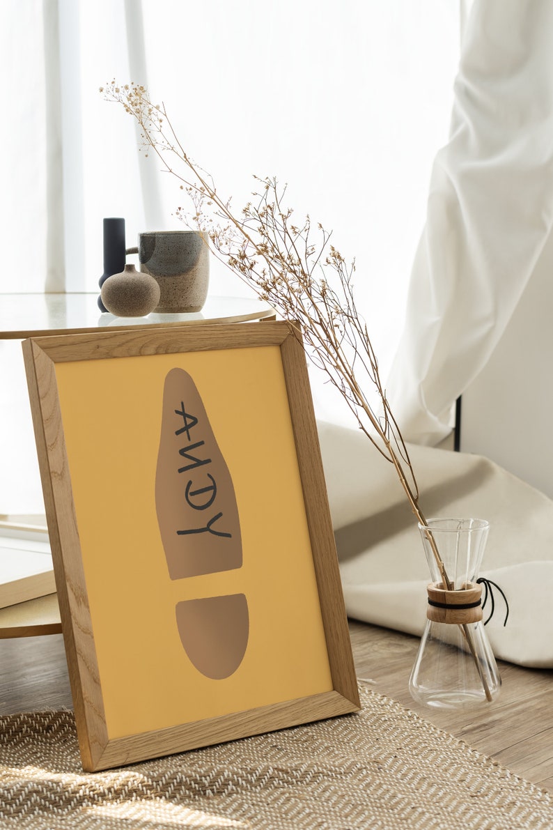 May include: A framed print with a mustard-yellow background features a brown footprint design with the name "ANDY" in black. A glass carafe with a wooden collar and dried flowers are also in the image. The frame is made of light-colored wood.