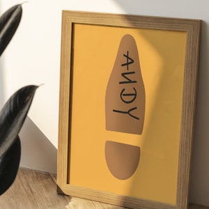 May include: A framed print featuring a brown footprint shape on a mustard yellow background. The word "ANDY" is written vertically in black within the footprint. The frame is made of light-colored wood, and the print is displayed on a wooden floor.