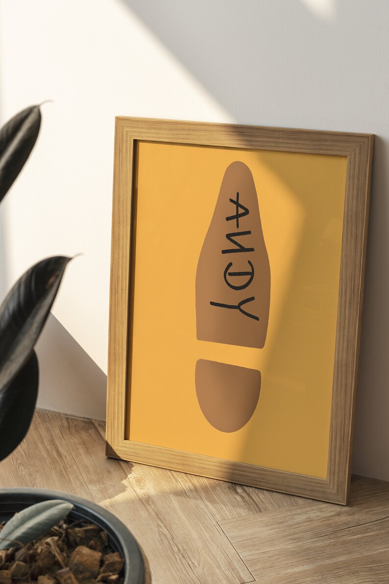May include: A framed print featuring a brown footprint shape on a mustard yellow background. The word "ANDY" is written vertically in black within the footprint. The frame is made of light-colored wood, and the print is displayed on a wooden floor.