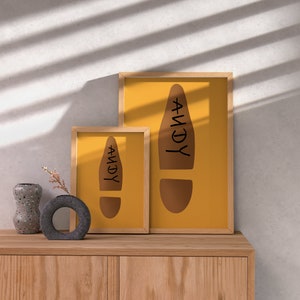 May include: Two framed prints with a mustard yellow background and a brown footprint design with the name "ANDY" in black. The frames are light wood. A decorative vase and a small round vase are on a wooden cabinet.