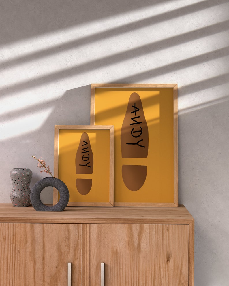 May include: Two framed prints with a mustard yellow background and a brown footprint design with the name "ANDY" in black. The frames are light wood. A decorative vase and a small round vase are on a wooden cabinet.