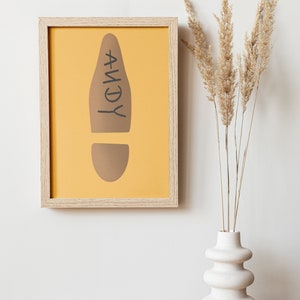 May include: A framed print featuring a brown footprint design on a yellow background, with the name "ANDY" in black. The frame is light wood. A white vase with dried plants is to the right.