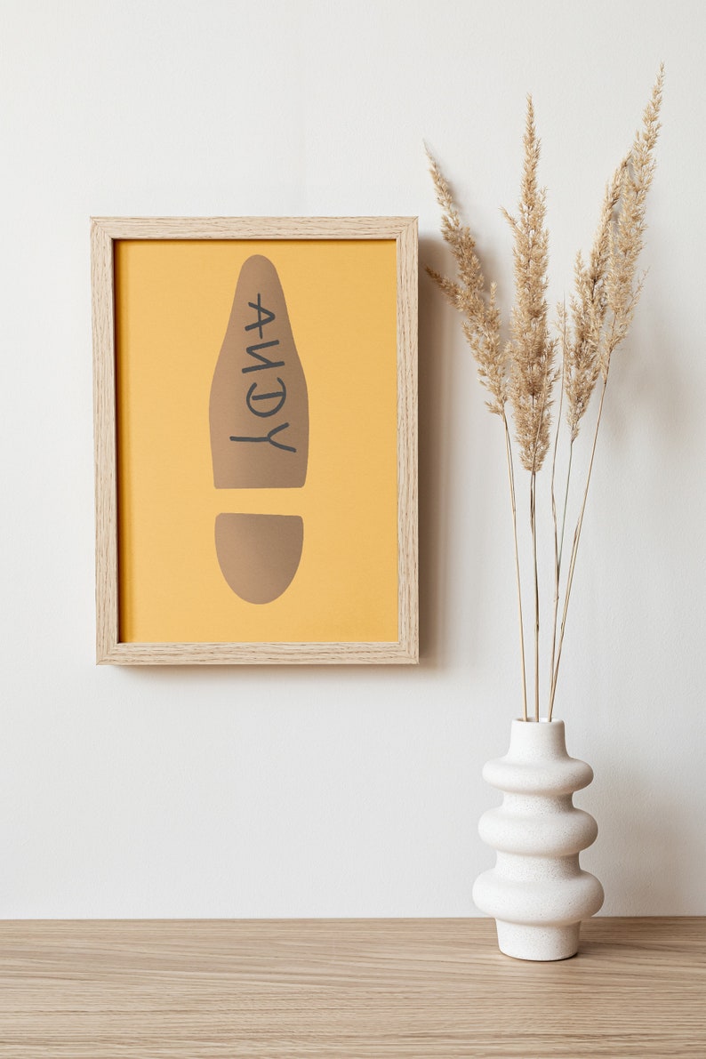 May include: A framed print featuring a brown footprint design on a yellow background, with the name "ANDY" in black. The frame is light wood. A white vase with dried plants is to the right.