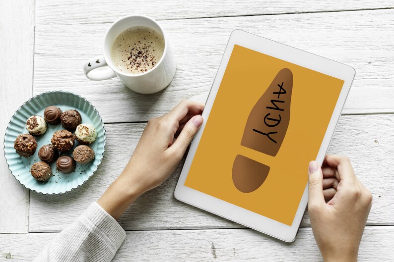 May include: A digital tablet displaying a brown footprint graphic with the name "ANDY" in black. A cup of coffee and a plate of assorted chocolates are on a white wooden surface.