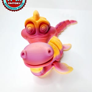 Könnte beinhalten: Ein 3D-gedrucktes, bewegliches Seeungeheuer-Spielzeug in Rosa, Gelb und Orange. Das Spielzeug hat ein lächelndes Gesicht, Flossen und einen langen, segmentierten Schwanz. Das Logo Flexi Factory Authorized Seller befindet sich oben links.