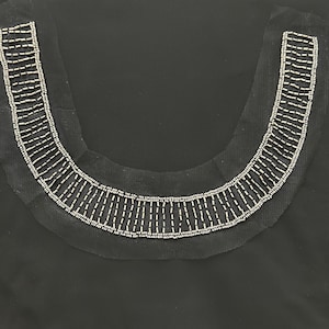 May include: Close-up of a black garment neckline embellished with silver-colored beads. The beads are arranged in a curved, symmetrical pattern, creating a decorative collar design. The fabric appears to be a sheer material.