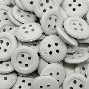 Può includere: Un mucchio di bottoni di plastica bianca con quattro fori ciascuno. I bottoni hanno un bordo rialzato e una superficie liscia. Alcuni dei bottoni hanno la parola "Prim" stampata su di essi.