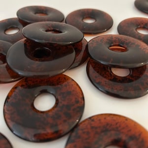 May include: Several glossy, dark brown, donut-shaped beads with a speckled pattern. Each bead has a central hole. They are displayed on a white surface. These beads are suitable for jewellery making or crafts.