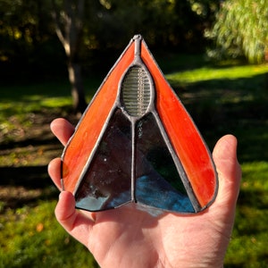 May include: A stained glass suncatcher in the shape of a heart with orange, blue, and black glass pieces. The suncatcher has a small oval-shaped window in the center.