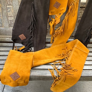 May include: A collection of suede chaps in shades of brown and yellow, with fringe detailing along the sides. Each chap features a small leather patch with text. The chaps are displayed on a wooden bench.