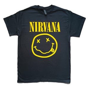 May include: A black short-sleeved t-shirt featuring the word NIRVANA printed in bold yellow capital letters above a yellow graphic of a smiley face with X-shaped eyes and a tongue sticking out from the side of a wavy, uneven mouth.