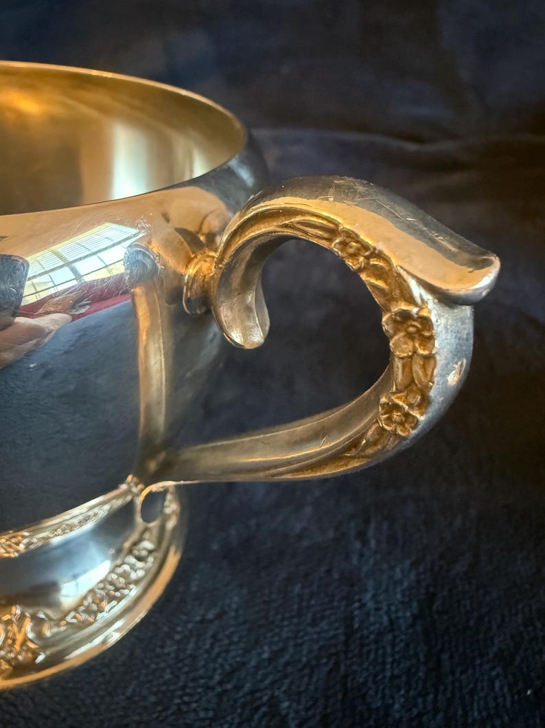 May include: A silver-colored cup with a decorative handle featuring floral detailing. The cup's interior reflects light, and the handle curves gracefully. The cup sits on a dark blue surface.