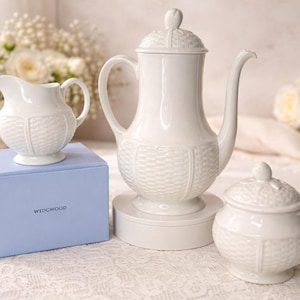 May include: A white ceramic tea set with a textured basketweave design. The set includes a teapot, creamer, sugar bowl, and a blue box with the word "WEDGWOOD" printed on it. The set is displayed on a white lace tablecloth.