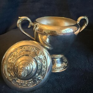 May include: A silver sugar bowl set with a lid and a small creamer. The sugar bowl has two ornate handles and a reflective surface. The lid is decorated with detailed engravings. The creamer has a matching design.