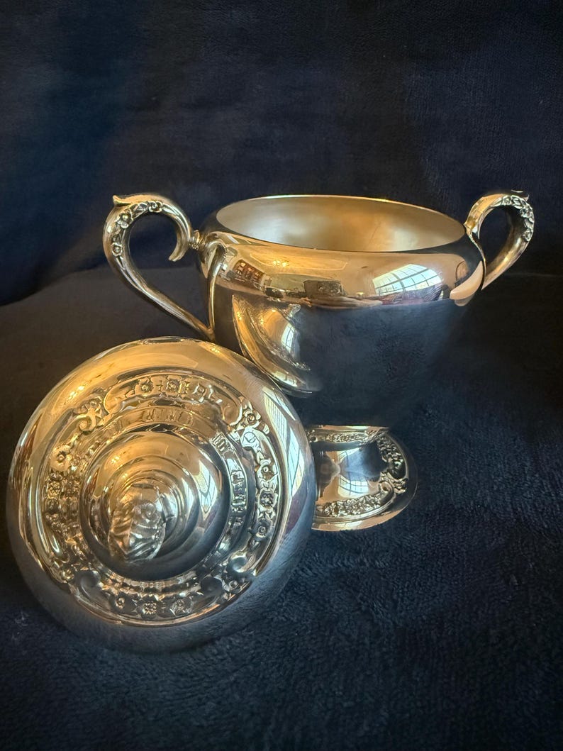 May include: A silver sugar bowl set with a lid and a small creamer. The sugar bowl has two ornate handles and a reflective surface. The lid is decorated with detailed engravings. The creamer has a matching design.