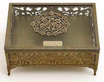 May include: An antique brass jewelry box with a rectangular shape and ornate floral detailing. The lid features a decorative floral oval and a small rectangular plaque. The box is raised on four small feet and has a clear glass top.