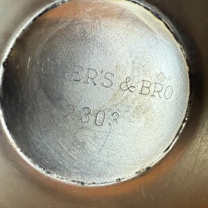 May include: Close-up of a silver-colored metal object with the text "ROGERS & BRO" and the number "2303" etched into the surface. The object has a circular shape with a slightly textured appearance.